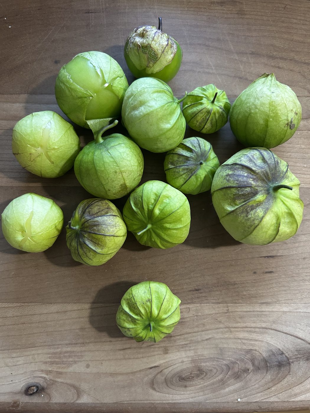 Tomatillos in their papery husks on a wooden board, pale green to yellow green with purple brown veining, two husks split to show the fruit inside